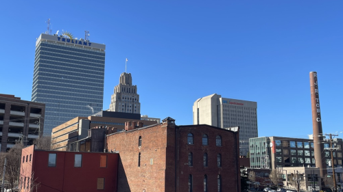 Brick buildings with smokestack and gray highrises in the background. A parking lot is full of cars in the bottom right corner.