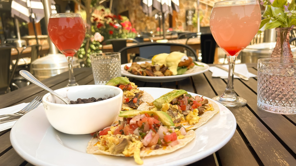 A plate of breakfast tacos topped with eggs and veggies sits on a brown wood table on the patio of The Mimosa House restaurant in Sacramento. Next to the plate, there are two pink-hued mimosas; across the table, there's another breakfast dish resembling a benedict.