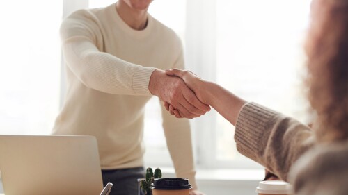 A person in a white sweater leans over a table to shake hands with a person in a beige sweater.
