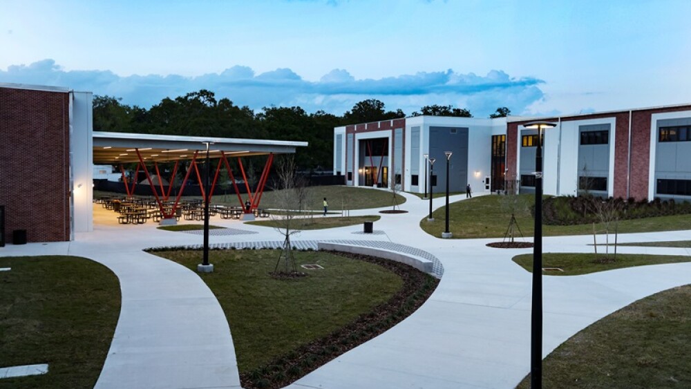 An outdoor courtyard at Southwest Middle School pictured at dusk. A two-story building and another building with an outdoor seating area are connected with curved sidewalks. Between the paths are light poles and newly planted trees.