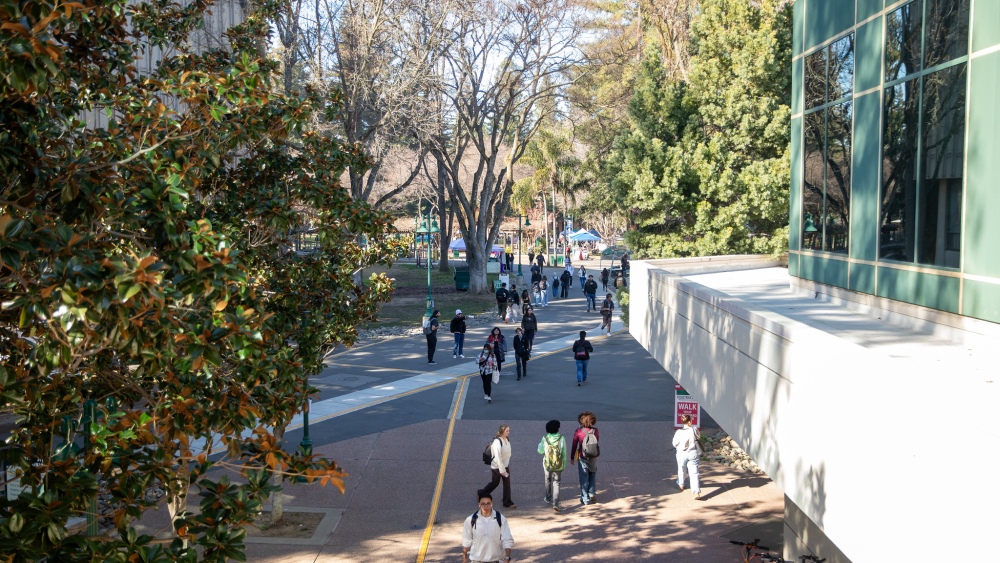 Students wearing backpacks walk between buildings at Sacramento State; large trees frame the campus.