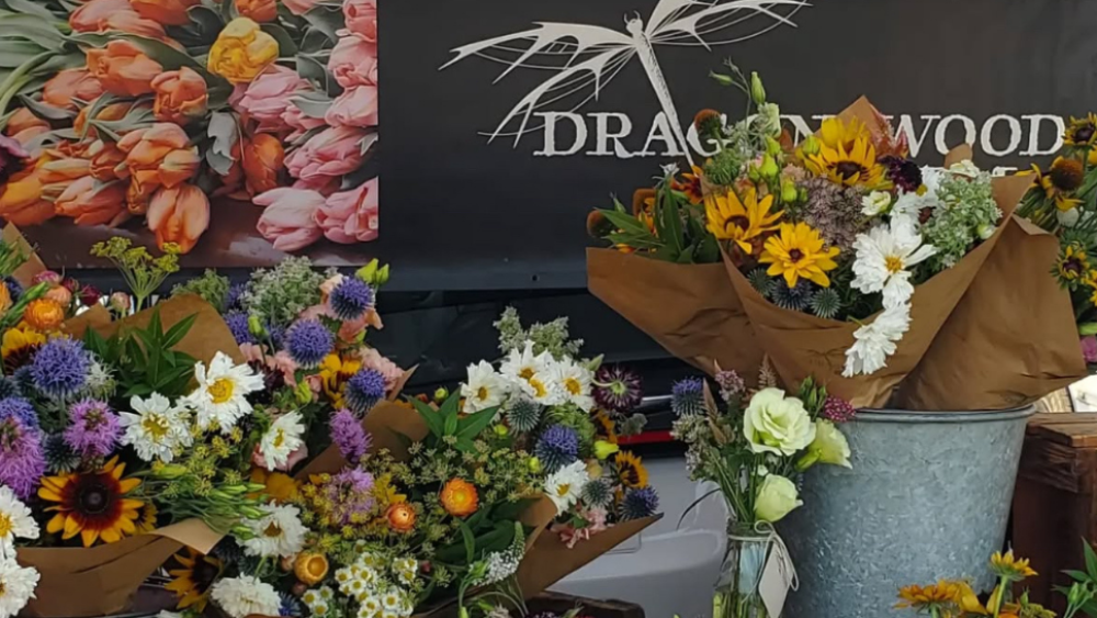 A black and white "Dragonwood" vendor sign with a dragonfly sketch poses behind brown paper-wrapped bouquets of white, purple, green, yellow, etc. flowers.