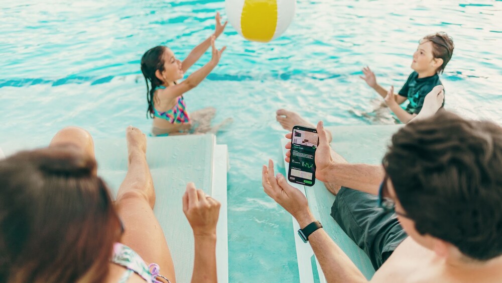 Two adults in lounge chairs look at rental properties on their phone while their kids play in the pool.