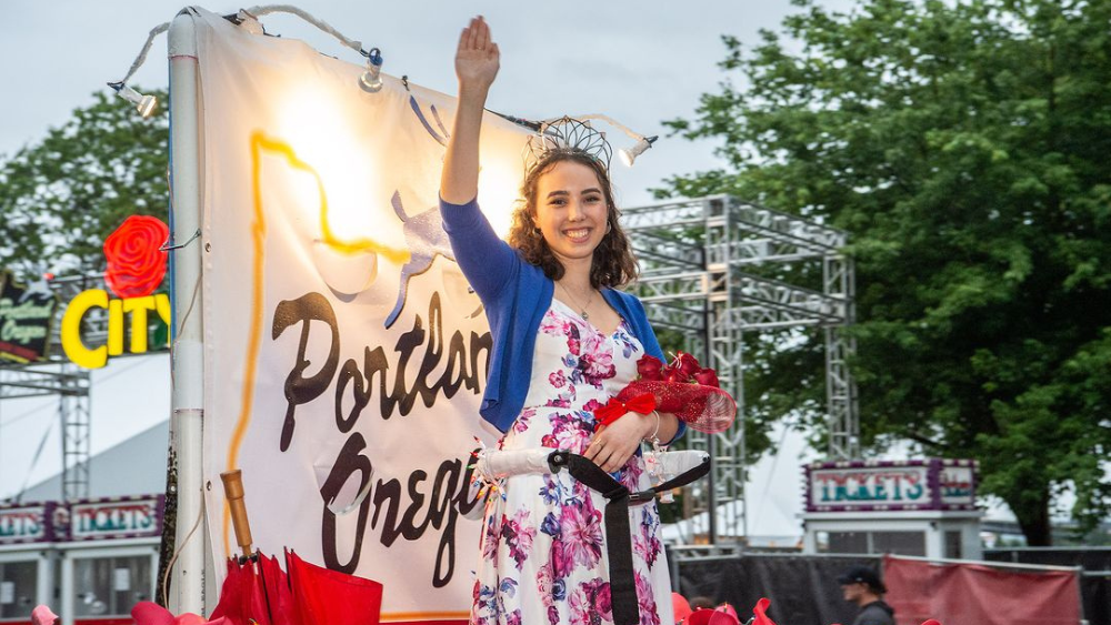 The Rose Festival Queen waves from a parade float that has the White Stag Sign as a background.