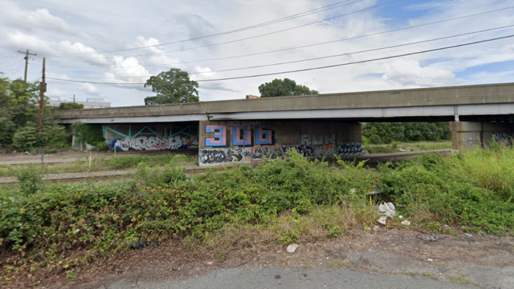 The image is a Google Streetview screenshot of the bridge of Arthur Ashe Boulevard. Graffiti marks the stone columns under the bridge, and litter scatters the grassy are in the forefront of the bridge.