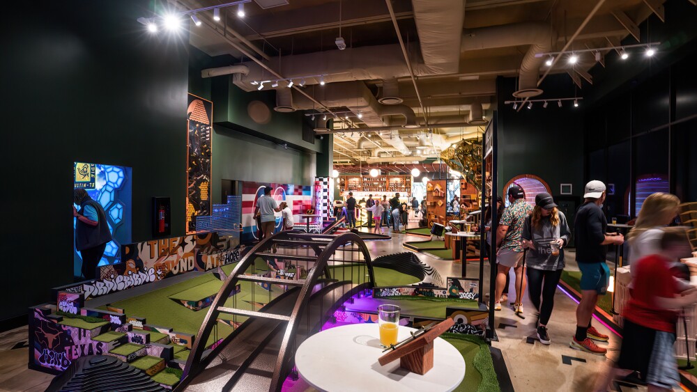 An indoor mini-golf course with various creatively designed holes and pathways. In the foreground, there's a small table with a drink and a golf scorecard. The mini-golf course features colorful and artistic elements, including graffiti-style decorations and unique structures like bridges and tunnels. Several people are seen playing mini-golf, adding to the lively atmosphere. The background includes a well-lit area with more mini-golf holes and a bar where people are gathered, enhancing the social and recreational ambiance of the space.