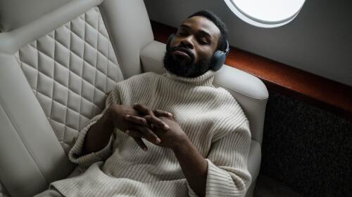 A man serenely resting on a flight by the window seat, with a cashmere sweater and headphones on.