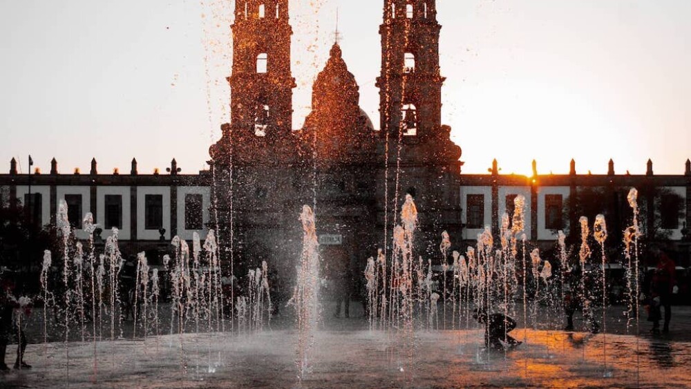 A sunset behind a two towered basilica and water fountains.