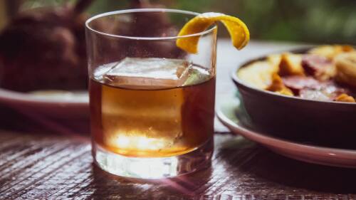 A glass of whiskey with a lemon peel garnish and a large ice cube.