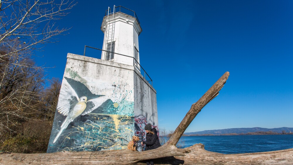 A dog and a woman wearing winter clothes sit on a fallen tree in front of a lighthouse by the Columbia River