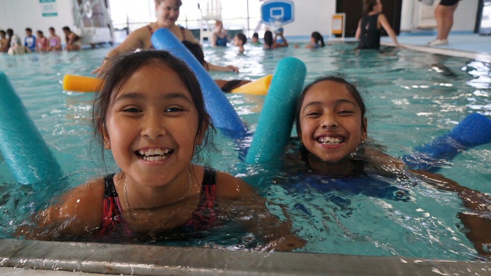 two kids floating on pool noodles in a pool smile