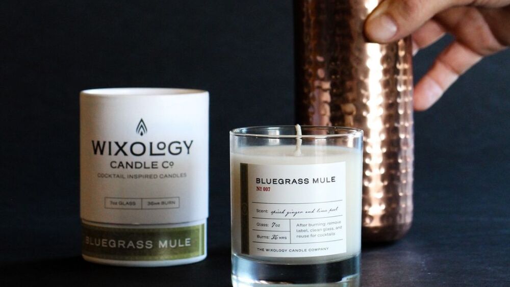 A photo of a hand holding a copper cocktail shaker next to a candle.