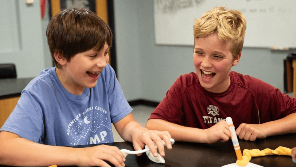 Two kids laugh at a table together as they work on an experiment or craft involving markers.