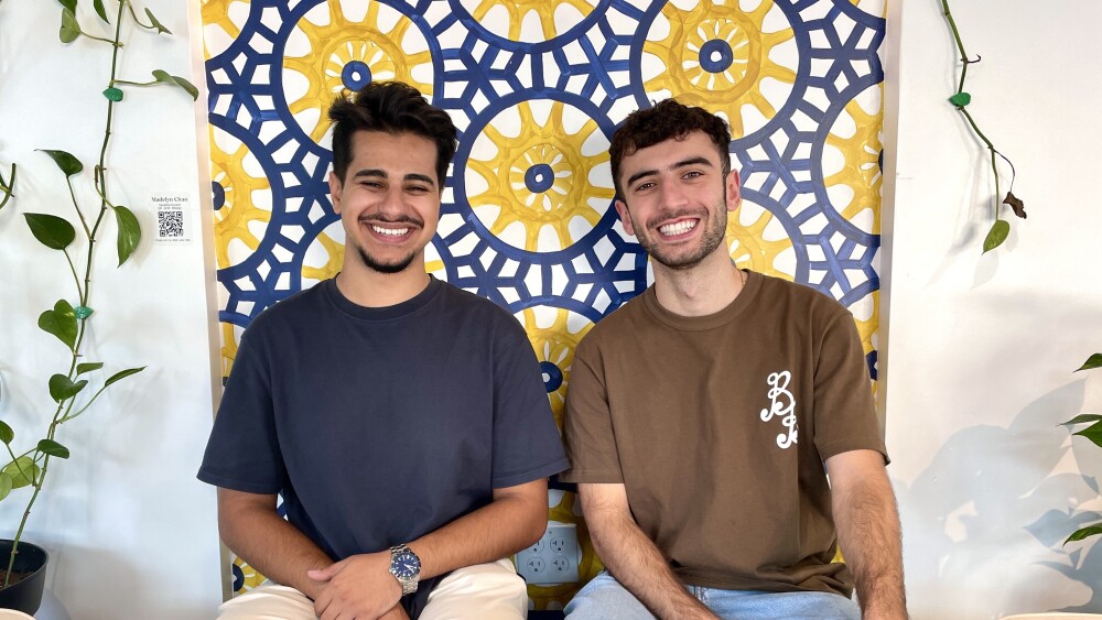 Toasted creators Jaafar Altameemi and Murat Akyuz sit shoulder to shoulder in front of a painted mosaic of blue and yellow geometrical patterns. They're both casually dressed and smiling.