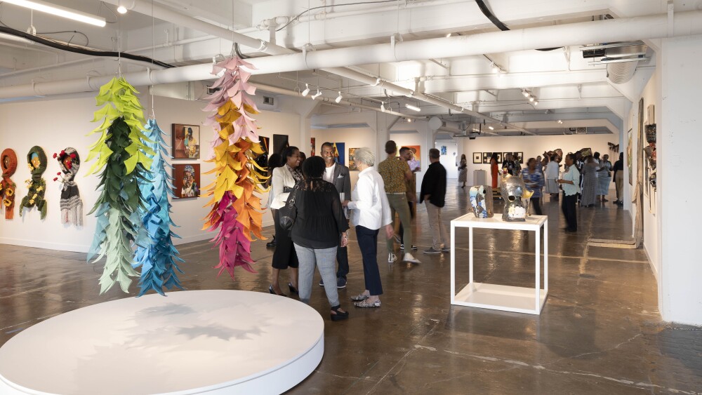 People stand in a gallery, some discussing in groups while others explore artwork around the room. A colorful leaf-like sculpture is suspended from the ceiling, featured prominently. Other displays include metal sculpture, textiles, and paintings.