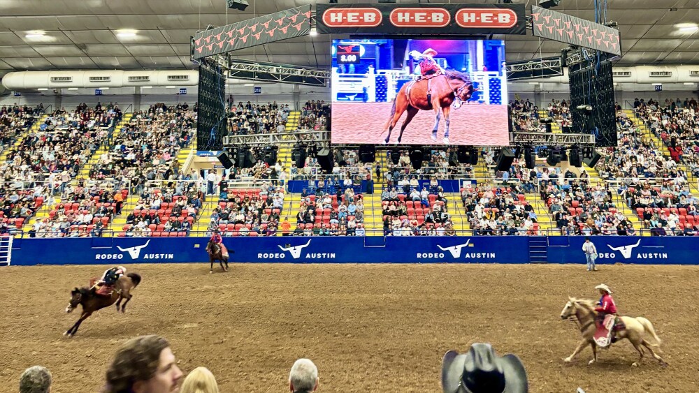 An indoor rodeo arena with a large crowd seated in multicolored stadium-style bleachers. The dirt arena floor features two horses and riders in motion, one wearing a red outfit and black hat. Above the arena is a large digital screen showing a close-up of a cowboy riding a bucking horse, with a H-E-B logo banner overhead. The ceiling displays a grid of bright lights and visible steel trusses. In the foreground, spectators are seated, including someone wearing a black cowboy hat.