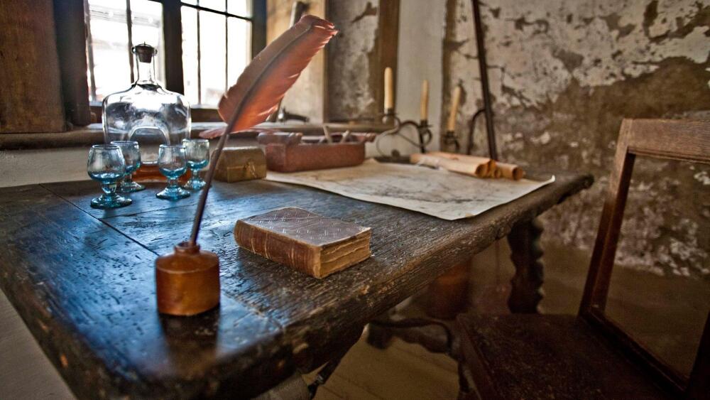 An interior shot of an old wooden table, chair, quill, book, and paper in front of a window.