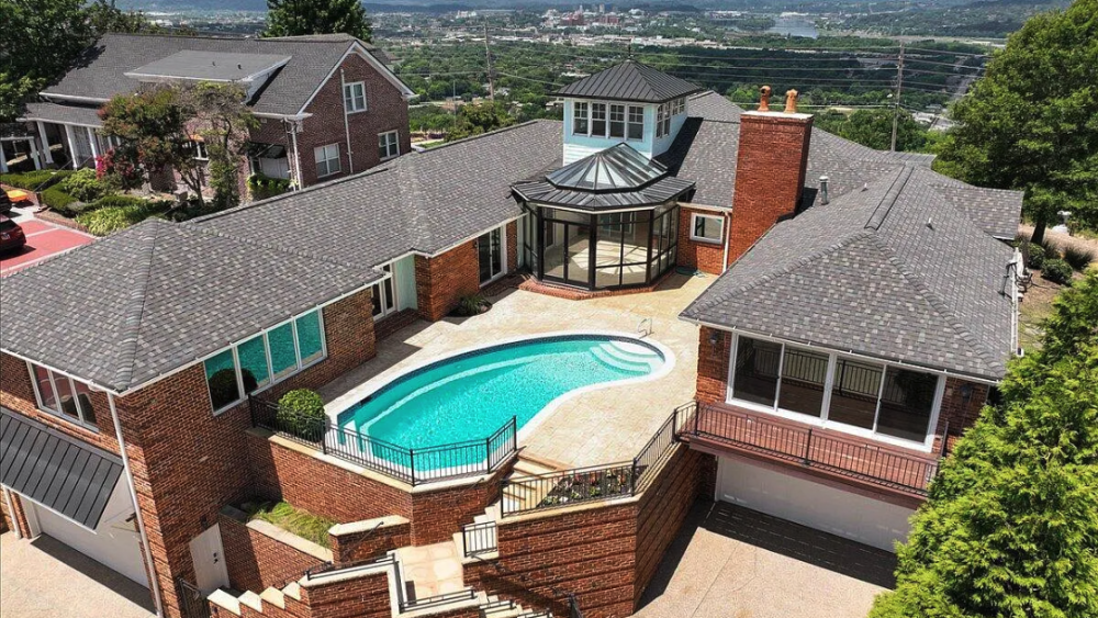 Aerial view of a luxurious brick house with a central glass-roofed sunroom and an oval swimming pool. Hilltop setting with scenic mountains.