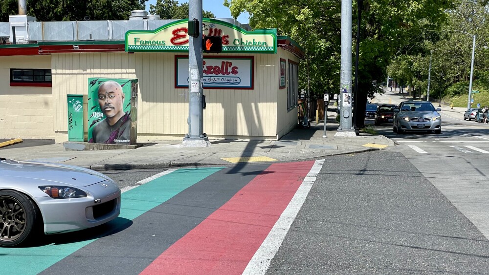 The crosswalk is painted in the red, black, and green stripe of the Pan-African flag. The crosswalk is leading one side of the street across to Ezell's Famous Chicken, a modest yellow building across from Garfield High School. 