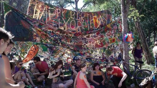 A group of people gather outdoors in a wooded area, sitting and chatting in front of a large, colorful, hand-painted banner that reads "Happy Birthday Eeyore." The banner is filled with vibrant, abstract art and patterns. Some people have body paint, and a decorated bicycle is leaned against a tree. The setting is festive and communal, with a creative, artsy vibe.