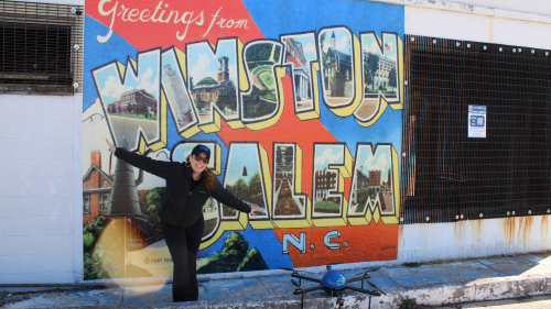 Woman in black jacket, pants, hat and sunglasses stands in front of a mural on a wall that says "Greetings from Winston-Salem, NC." Her arms are outstretched and a drone is on the sidewalk.