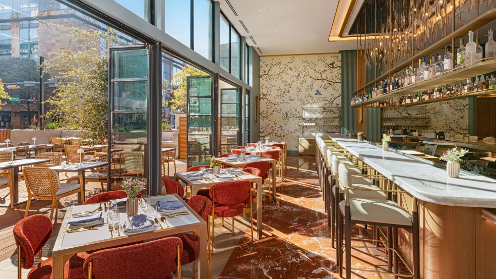 Bright and airy restaurant interior with floor-to-ceiling windows that open to an outdoor patio area. The indoor space features a marble-topped bar with elegant white stools, gold accents, and shelves lined with bottles and decorative lighting. Tables are set with white linens, gold cutlery, and vases of fresh flowers, surrounded by red upholstered chairs with brass details. A floral-patterned feature wall adds a touch of sophistication, while the outdoor area includes woven chairs and tables under sunny skies, creating a seamless blend of indoor and outdoor dining.