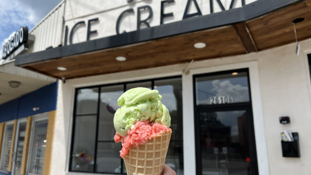 A hand holding up a waffle ice cream cone with two scoops of ice cream, one bright green (pistachio) and the other bright pink (watermelon).