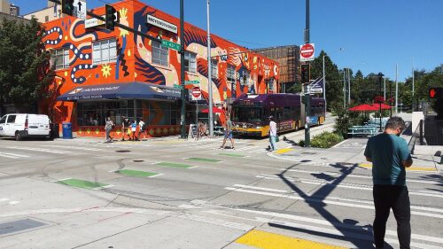 Pedestrians cross the street in the University District while a bus prepares to turn the corner. There's a bright red mural painted by Stevie Shao on one of the buildings in the background.