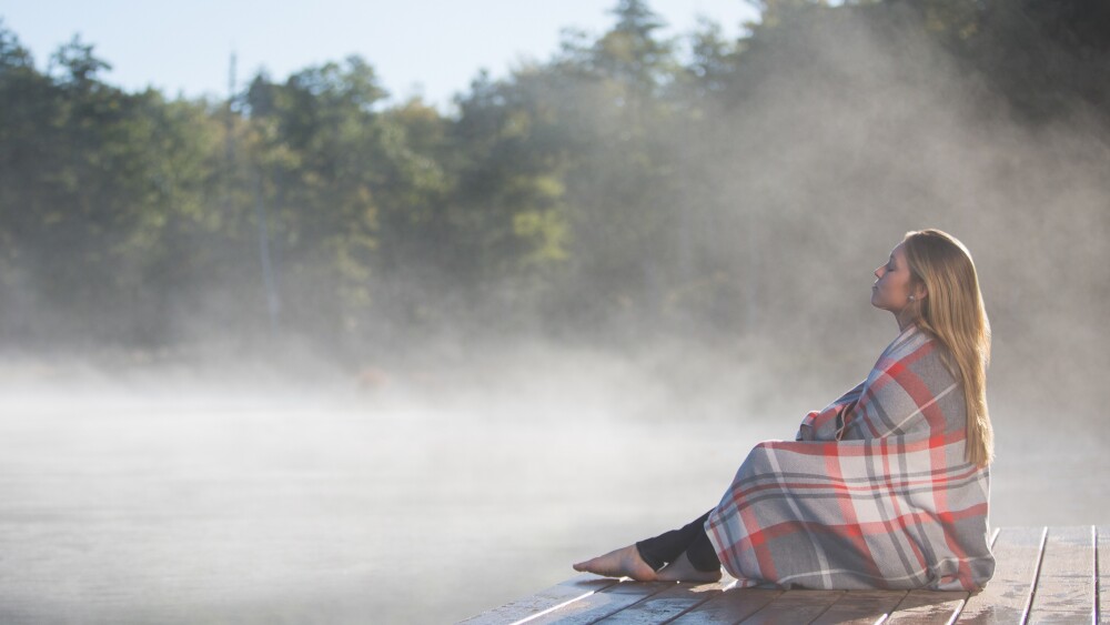 A woman wrapped up in a plaid blanket sits on a dock amongst mist on a lake.