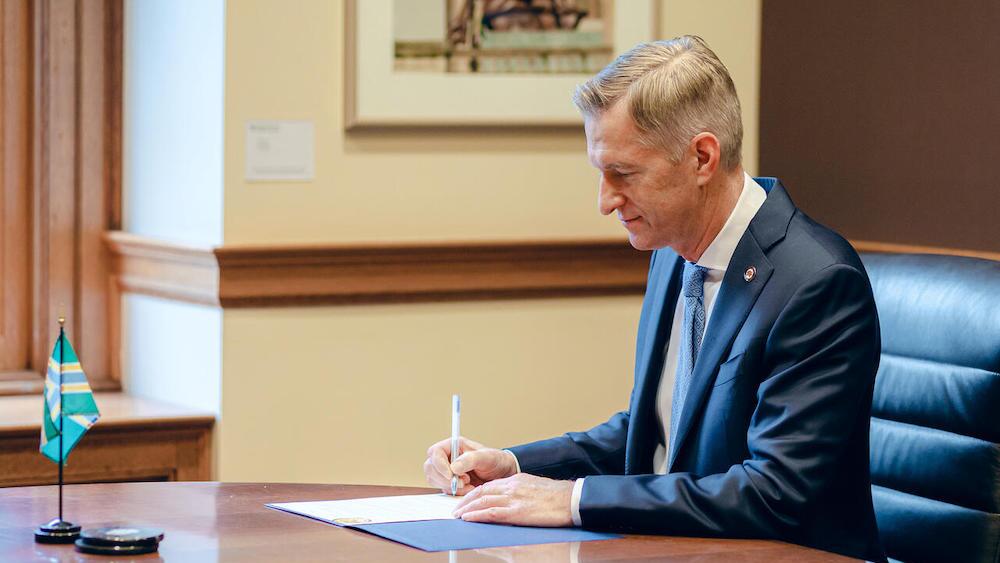 Portland Mayor Ted Wheeler signs a document sitting at a large desk with a small Portland flag on it.