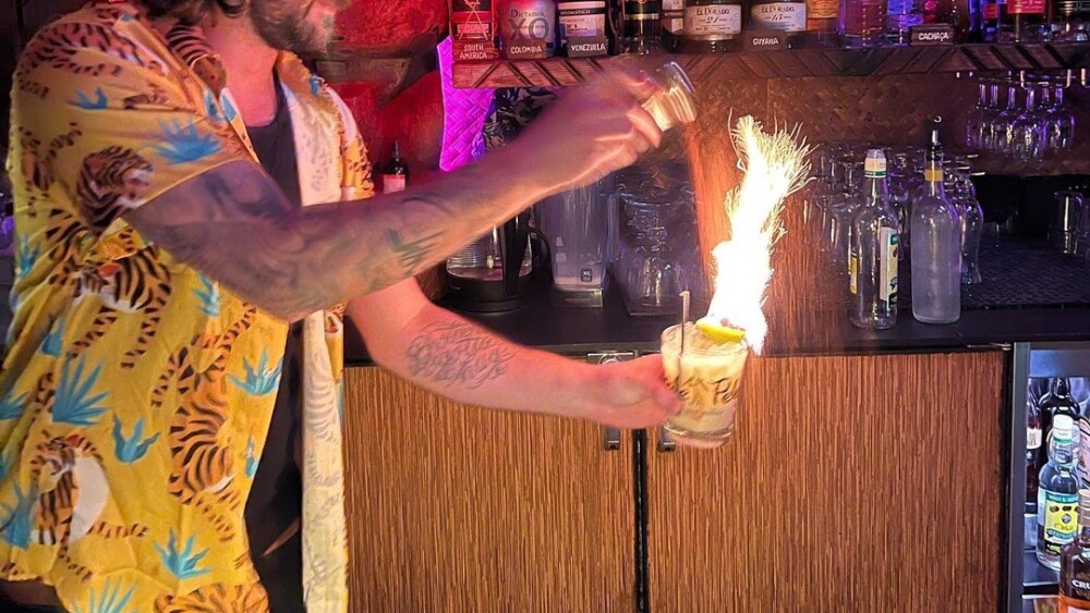 A bartender wearing a tropical shirt holds a flaming cocktail inside of a tiki bar.